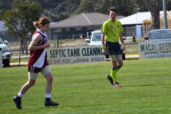 An umpire looks on as a female footballer jogs past.